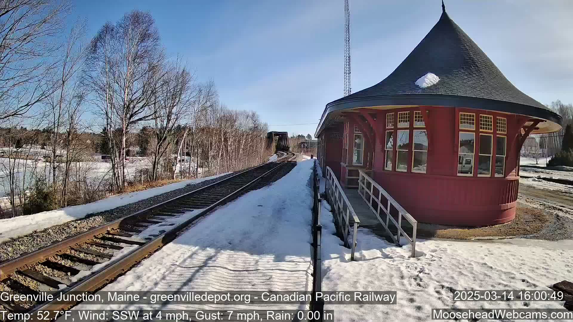 A red, round train station with a conical roof sits next to train tracks partially covered in snow under a clear blue sky.