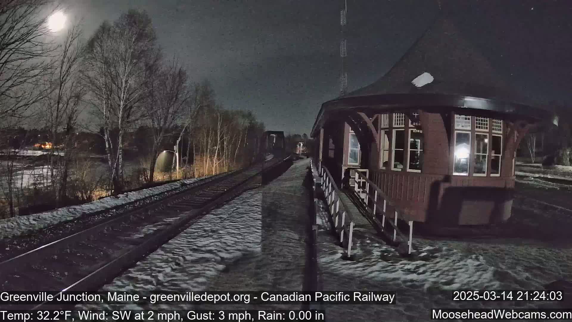 A nighttime view of a snow-covered train platform and tracks under a bright moon, with a dark-colored train station building visible to the right.