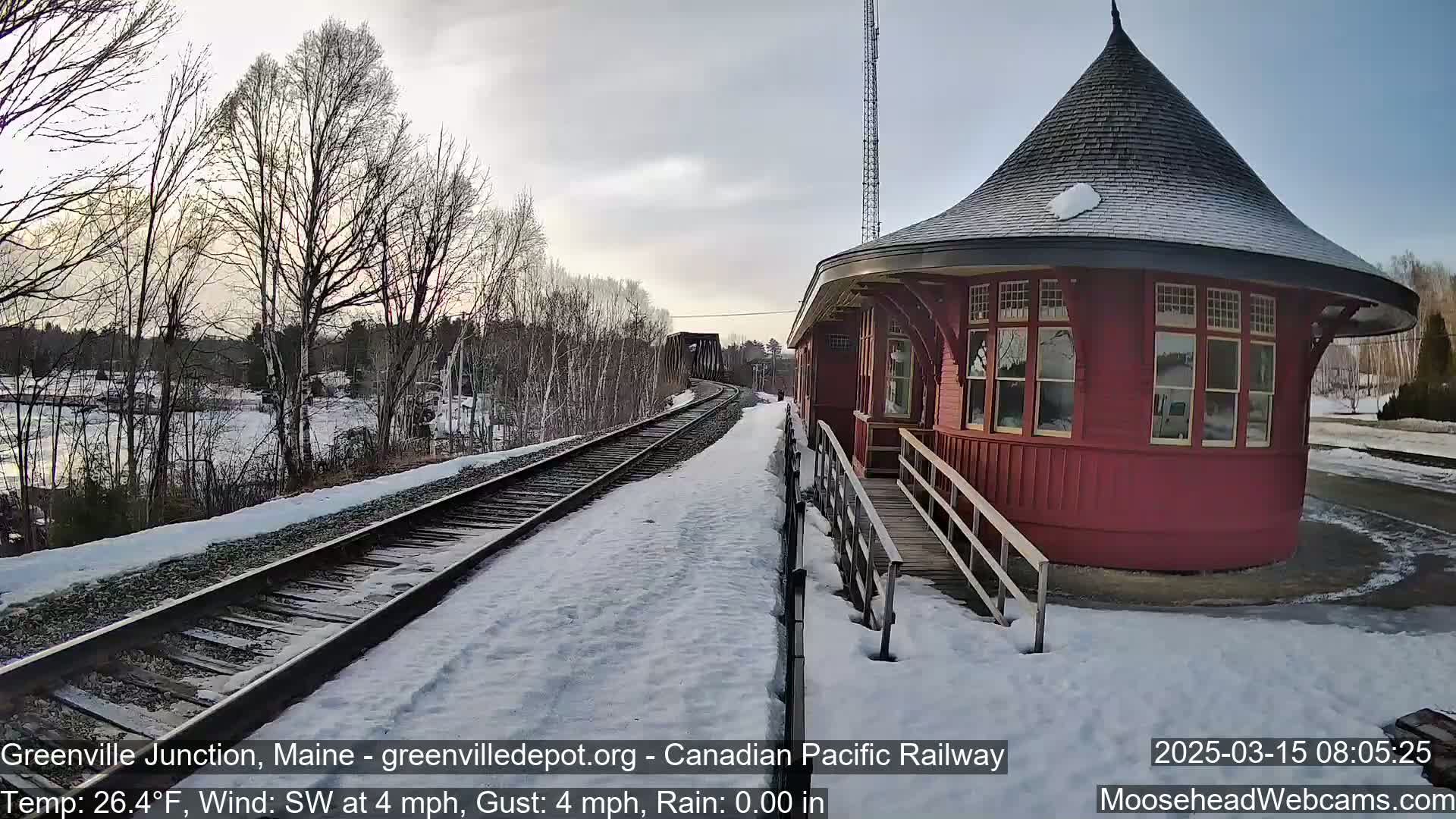A red, round train depot sits beside snow-covered railroad tracks on a partly sunny, cold day.