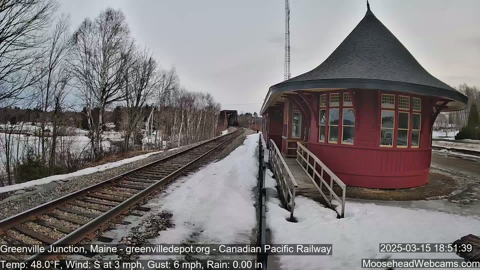 A red, round train station with a conical roof sits beside a snow-covered railway track on a cloudy day.