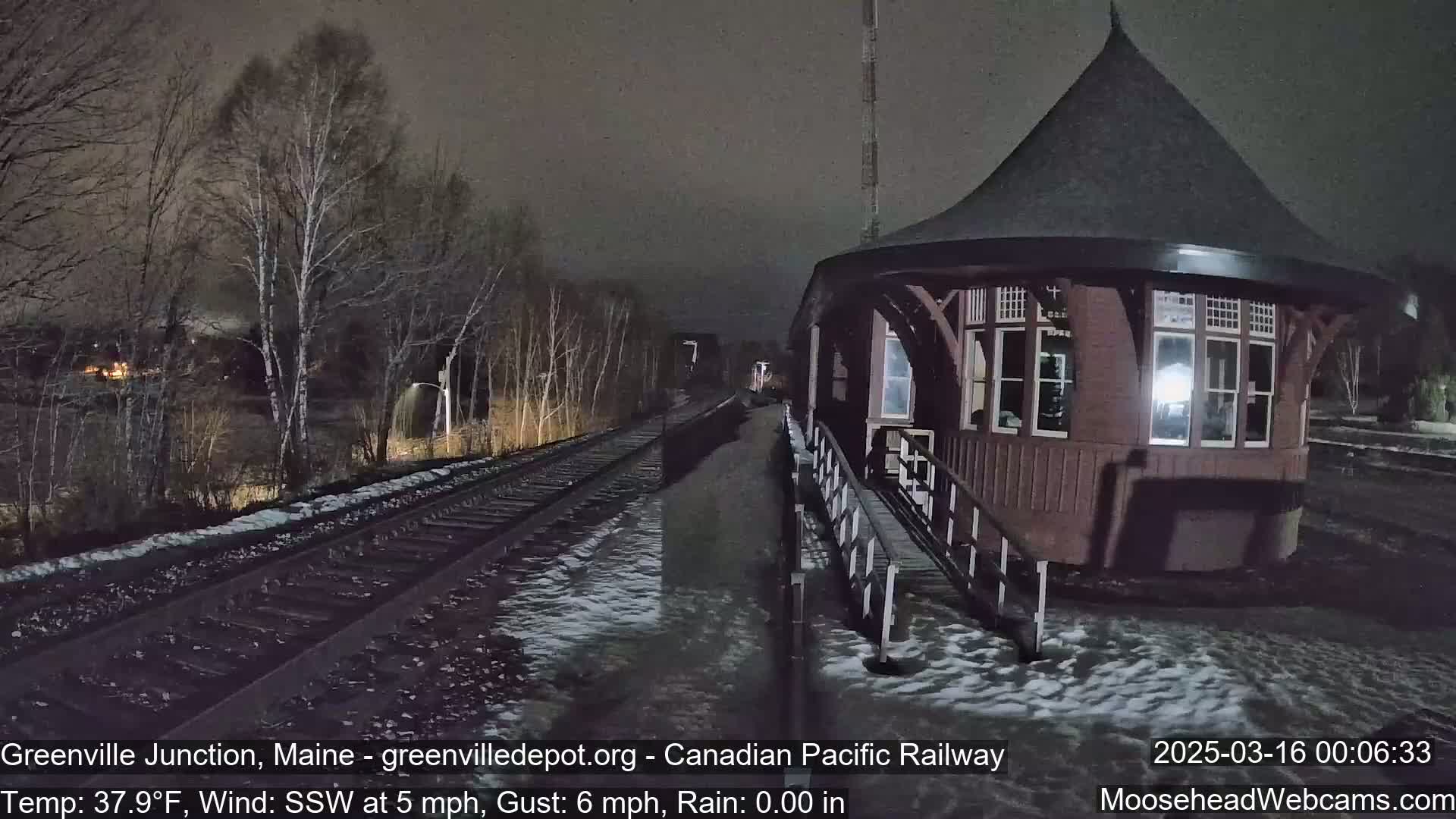 A dark, rainy night scene shows a train station with a round, wooden structure next to railroad tracks, surrounded by bare trees under a mostly dark sky.