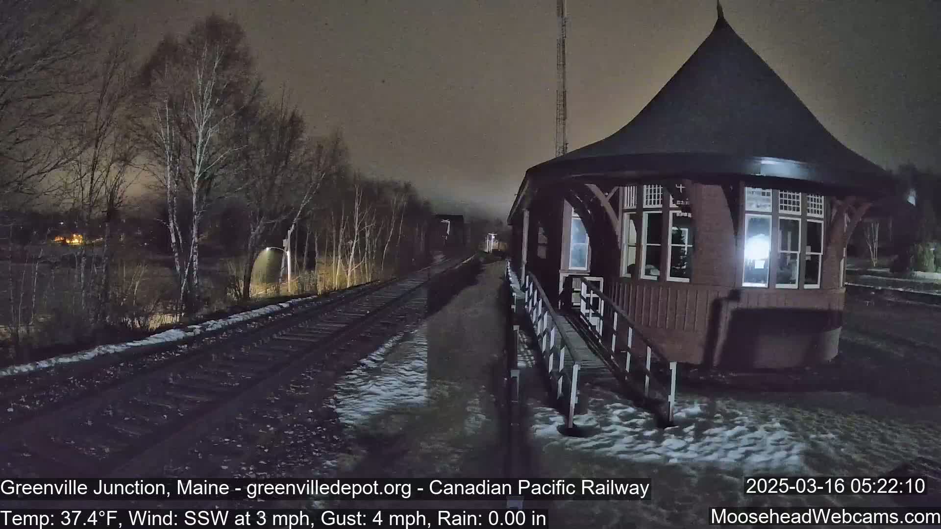 A dark, nighttime view of a train depot and railway tracks with light snow on the ground and a temperature of 37.4°F, a south-southwesterly wind, and no rain.