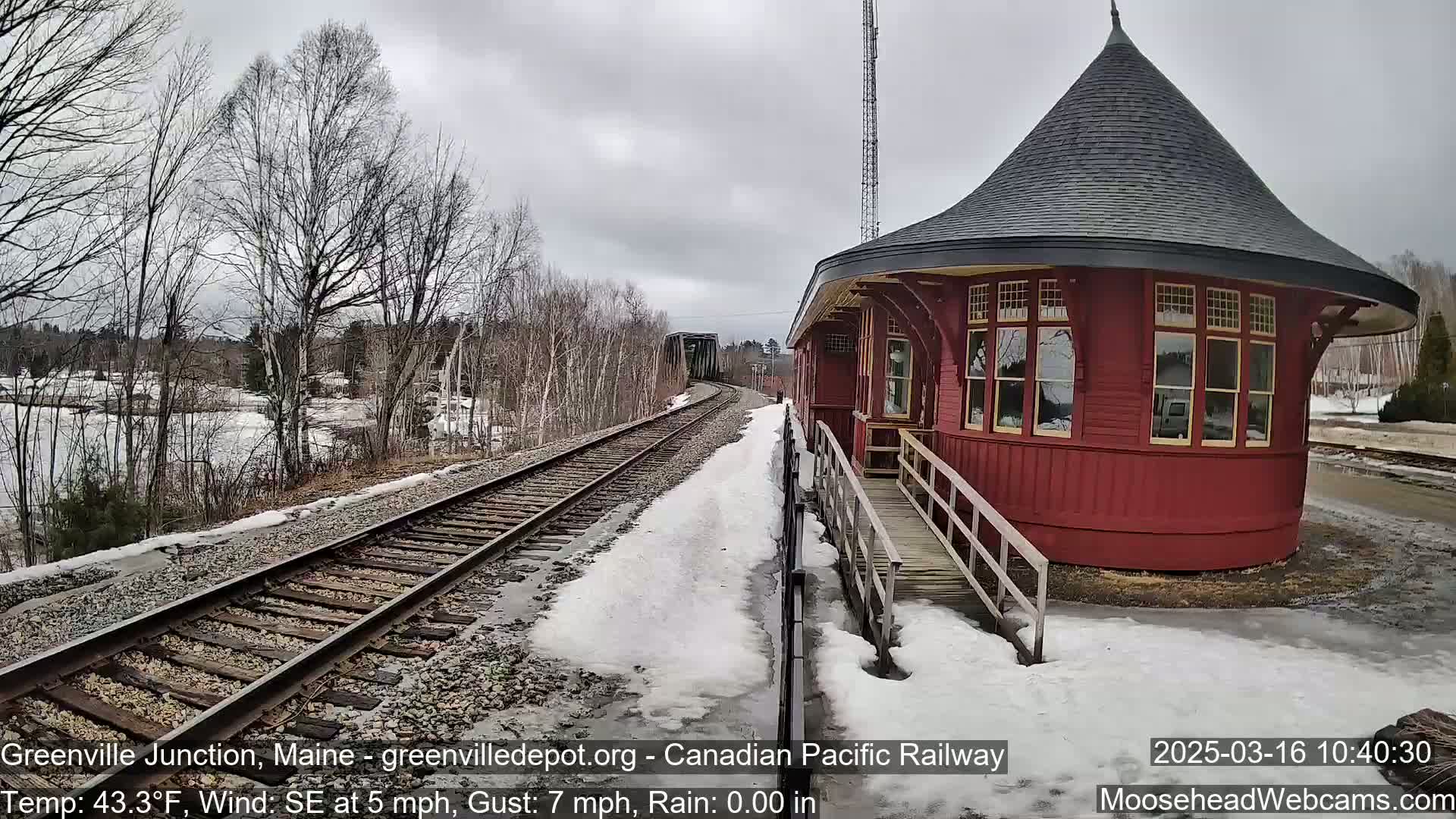 A red, round train depot with a conical roof stands beside a railway track on a partly snowy, overcast day.