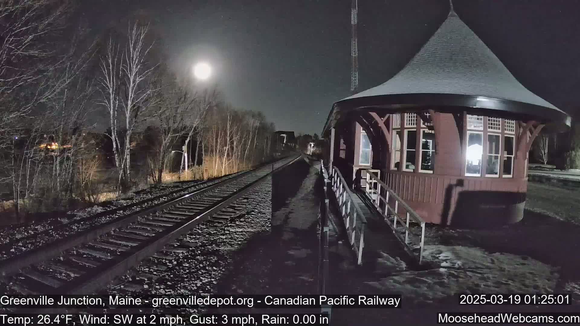 A brightly lit train station stands beside railroad tracks under a nighttime sky with a full moon, light snowfall on the ground, and a southwest wind.
