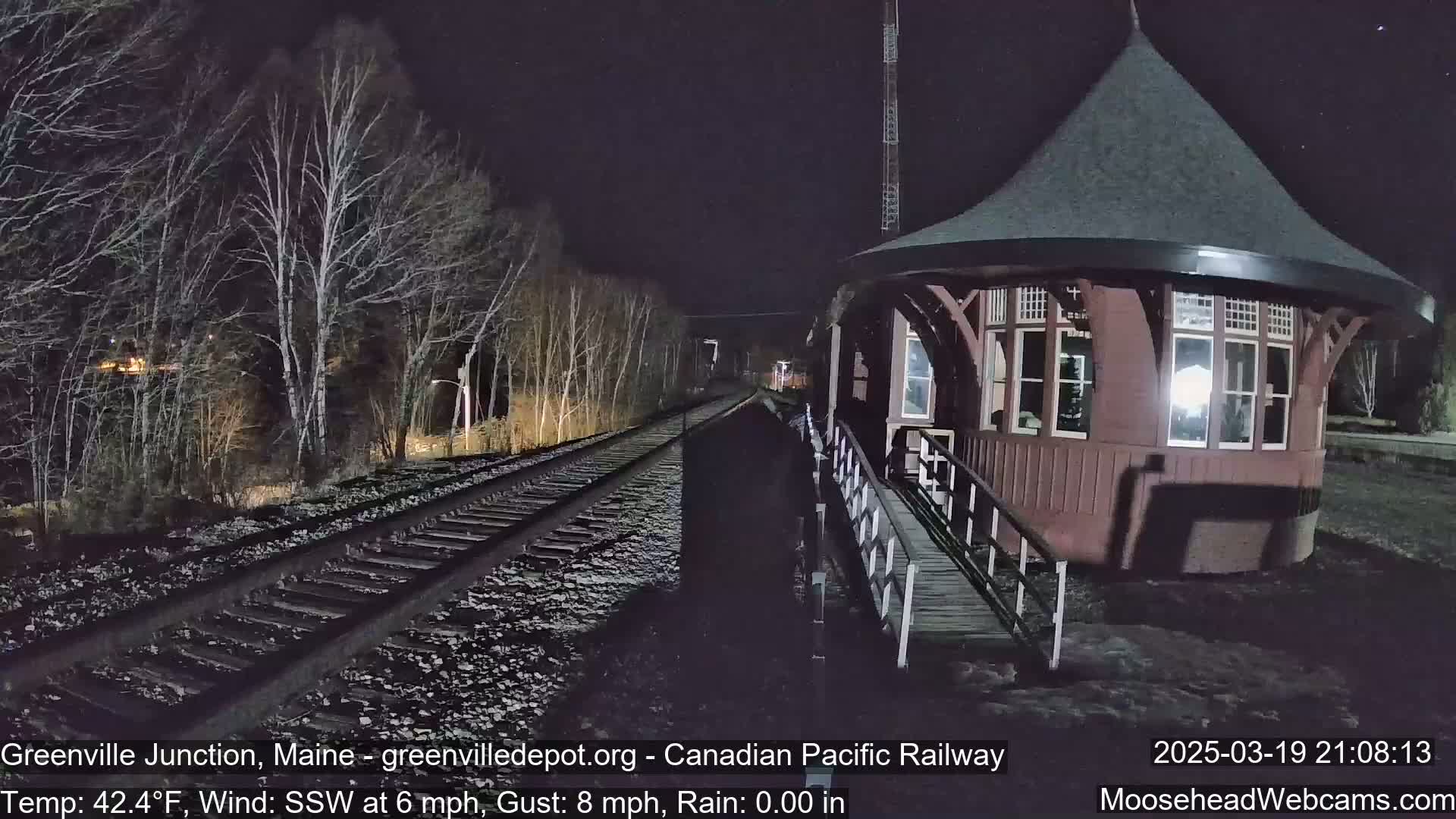 A train track stretches into the night past a round, dark-red train station under rainy conditions.