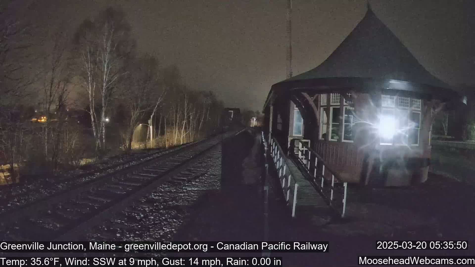 A nighttime view of a train station and railroad tracks under a dark, rainy sky.