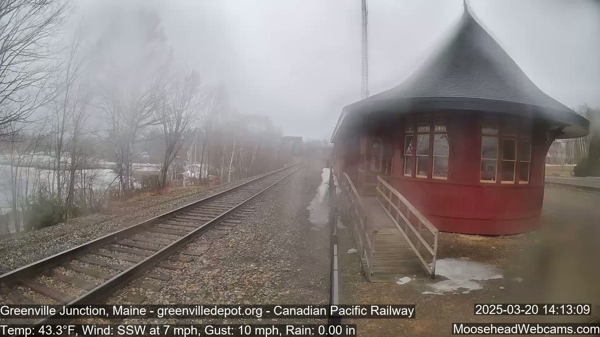 A red, round train depot is visible beside railroad tracks on an overcast, rainy day.