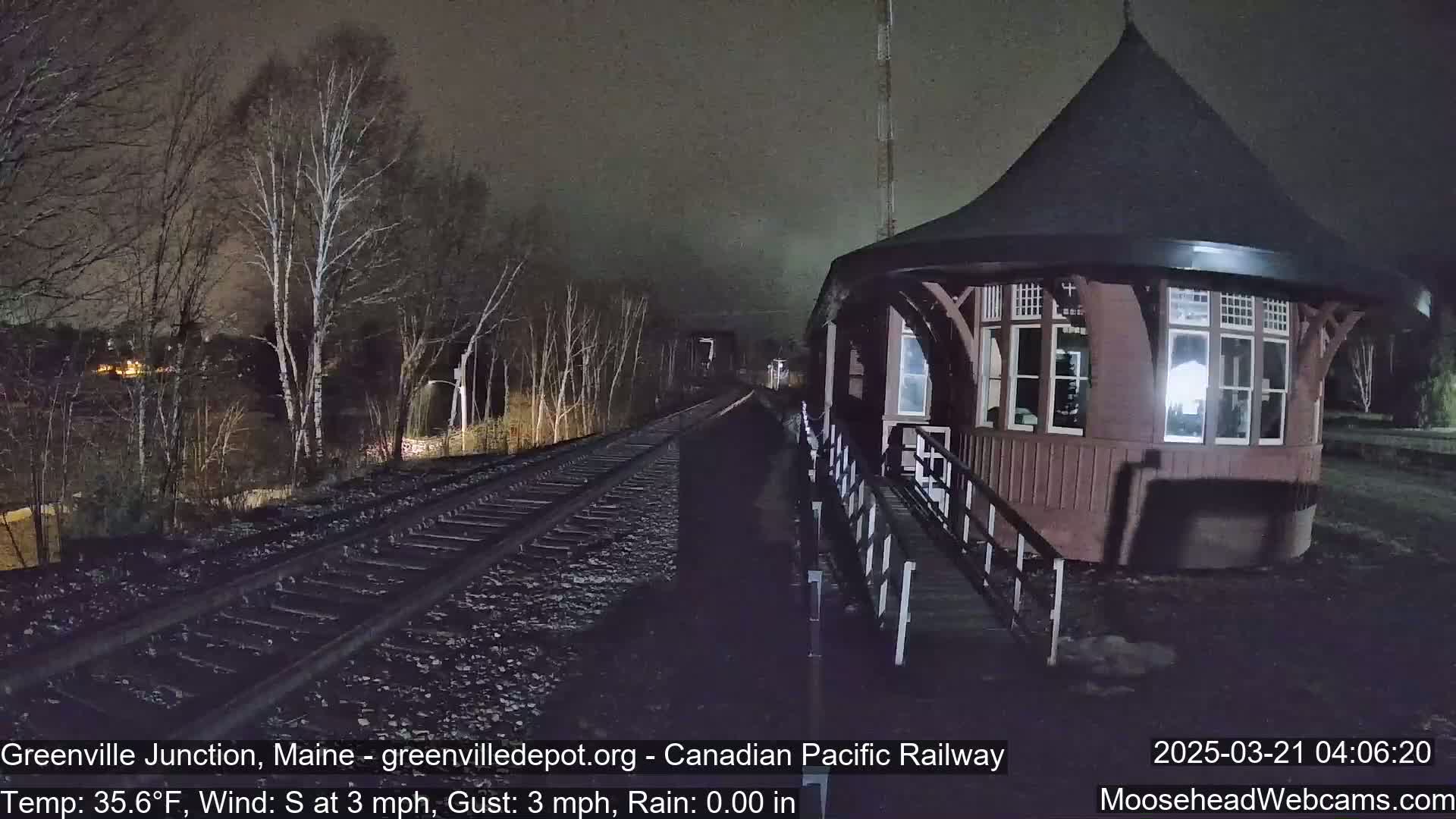 A dark brown, round train depot with a peaked roof stands beside train tracks at night under a clear sky.