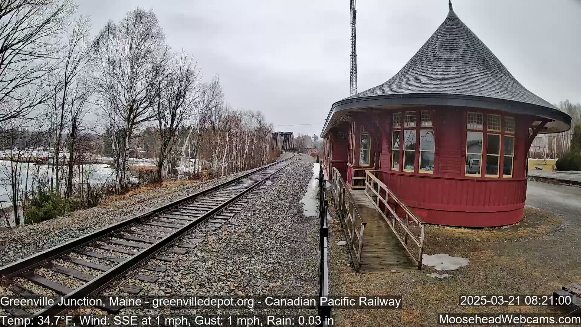 A red, round train station with a conical roof sits beside railroad tracks, under a gray, rainy sky.