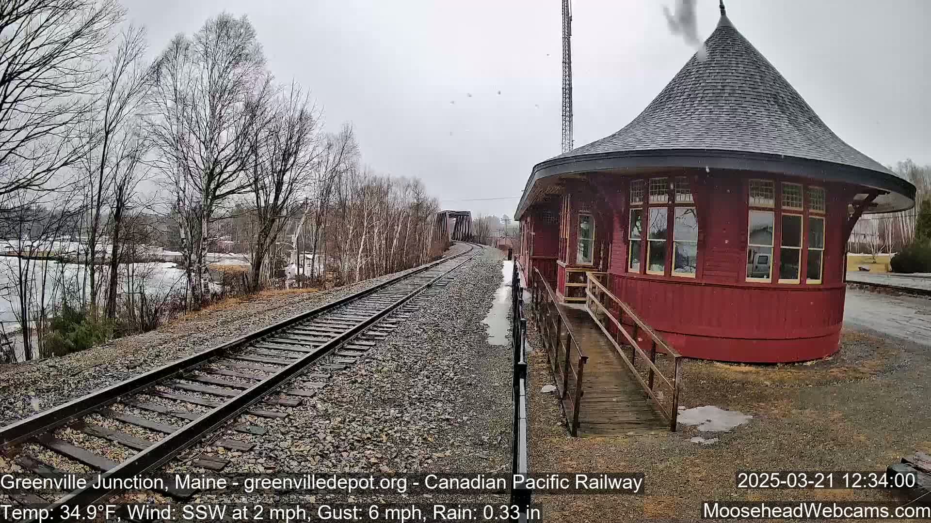 A red, round train depot stands beside railway tracks in light rain and snow, with bare trees and a body of water visible in the background.