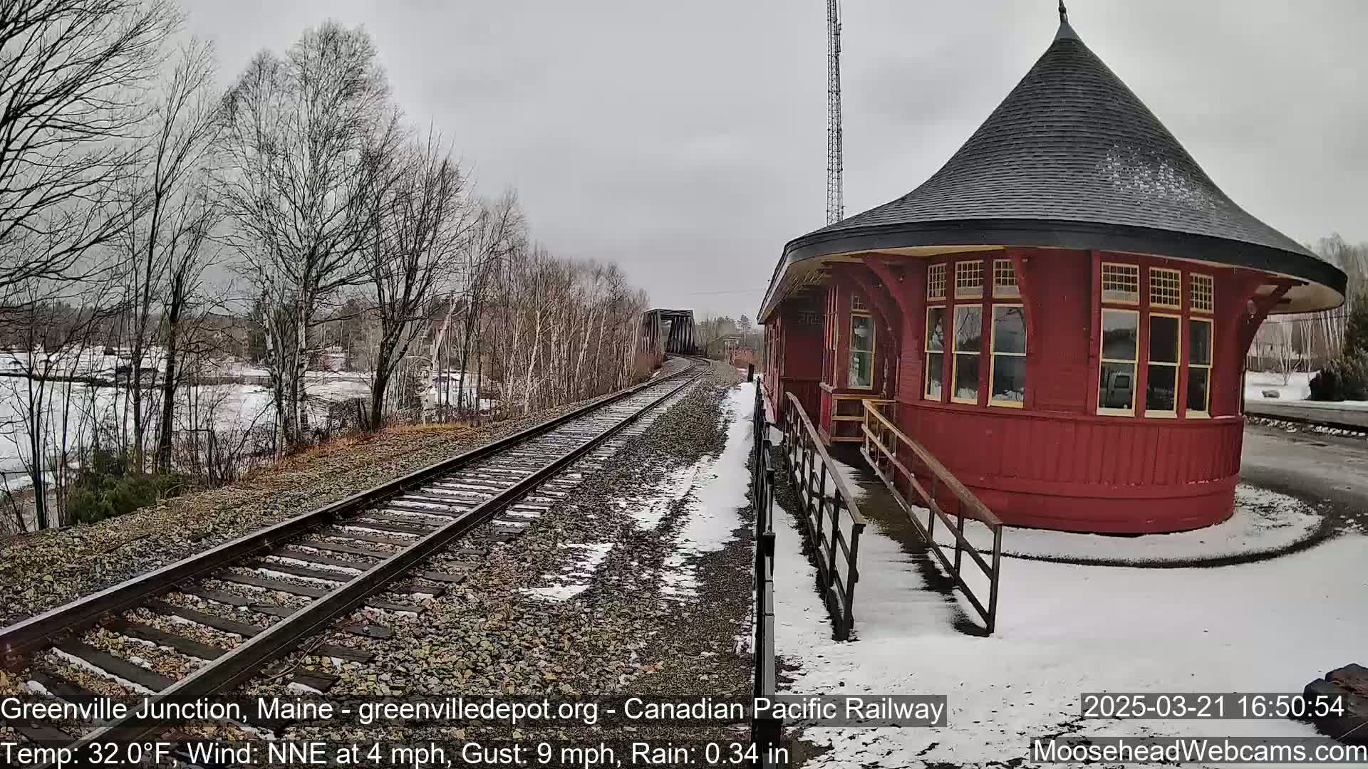 A red, round train station stands beside railroad tracks that stretch towards a bridge under a gray, rainy sky with patchy snow on the ground.