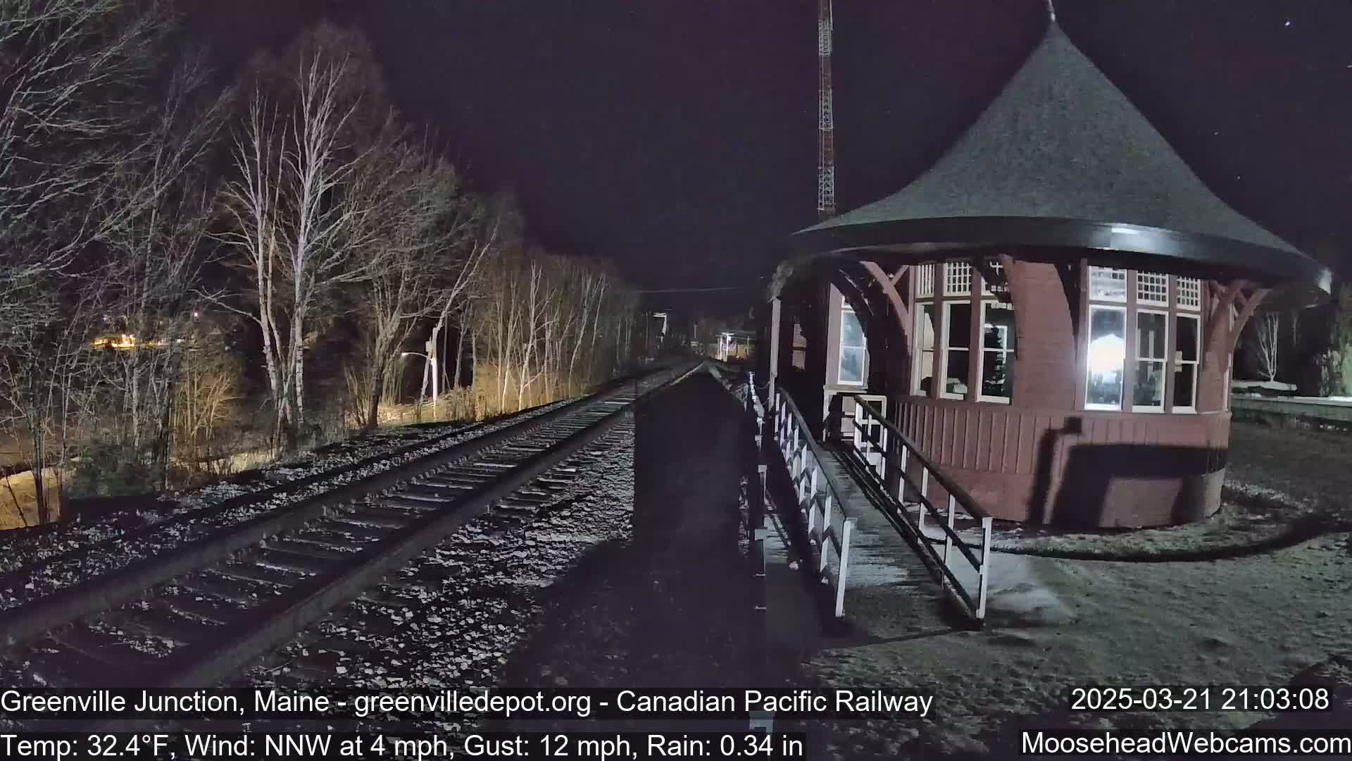 A dark, rainy night scene shows a round train station next to railroad tracks that extend into a bare, wooded area.