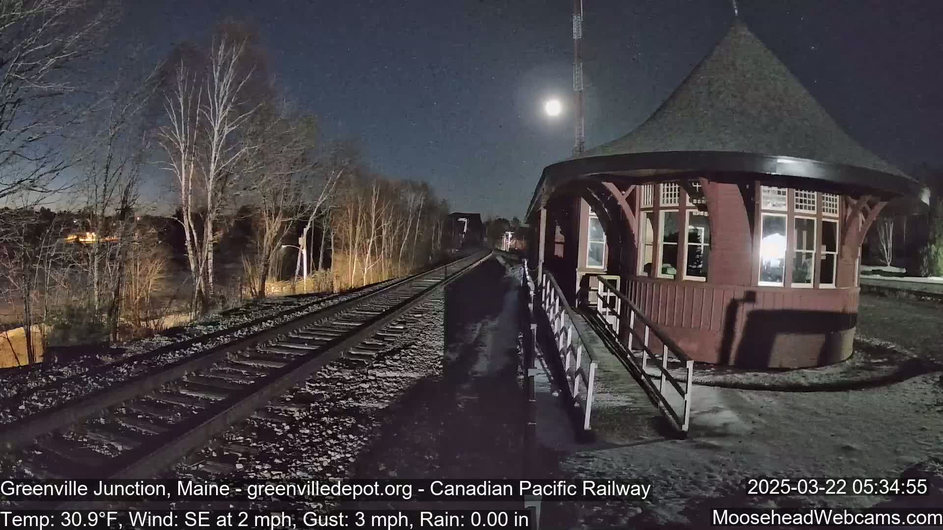 A snow-covered train platform with a round, dark-red building under a nighttime sky illuminated by a full moon.