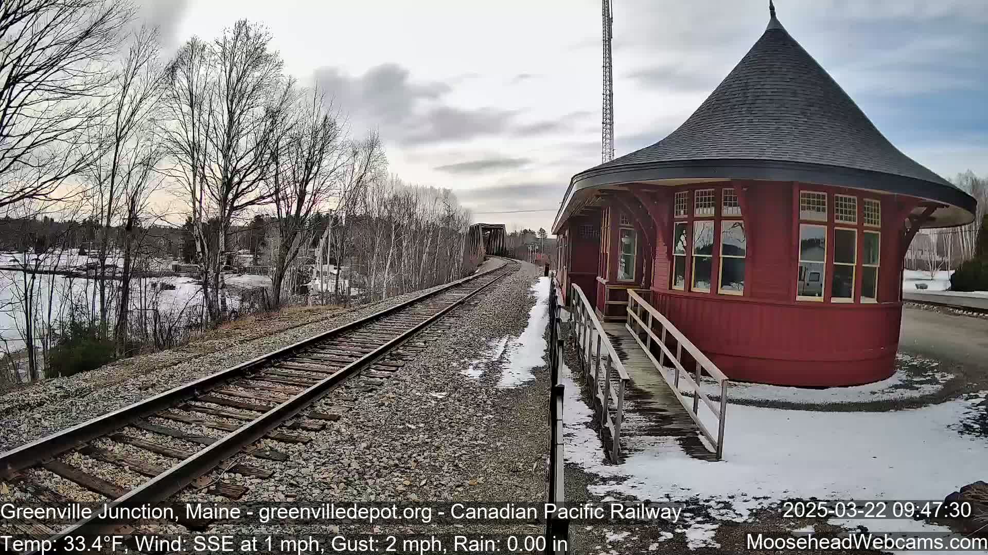 A red, octagonal train station stands beside a railway track curving towards a bridge, with a partly frozen body of water and bare trees visible in the background under a cloudy sky.