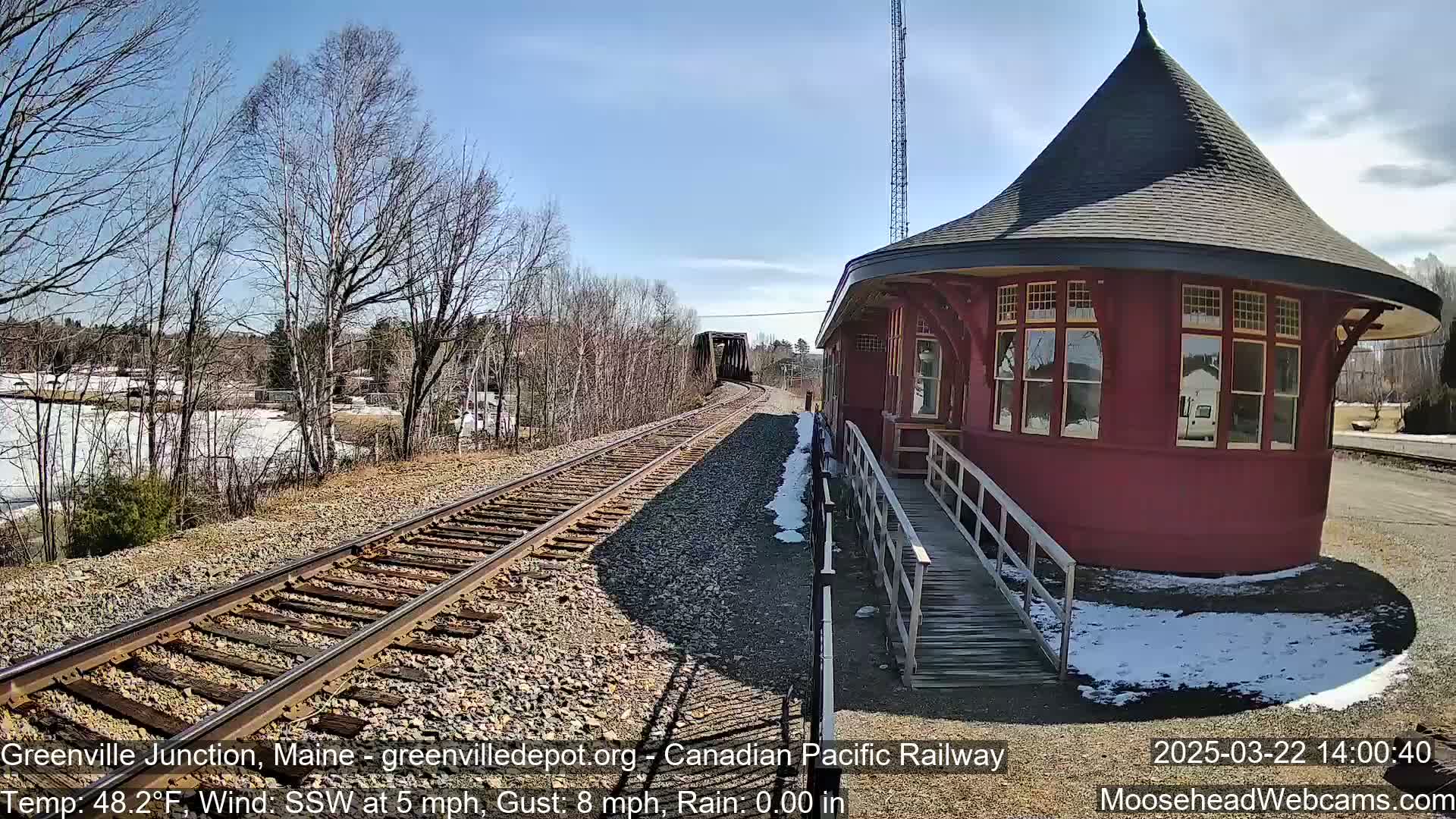 A red, round train depot with a ramp sits next to railroad tracks on a sunny day with patches of snow on the ground.