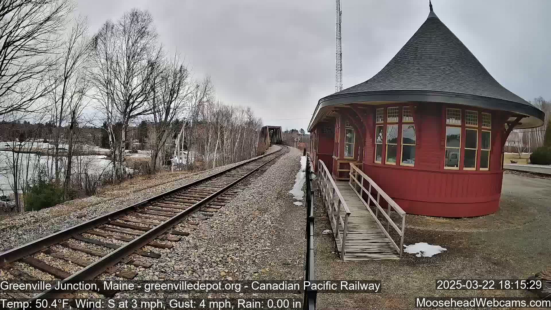 A red, round train depot with a conical roof sits beside train tracks curving towards a bridge under an overcast sky.