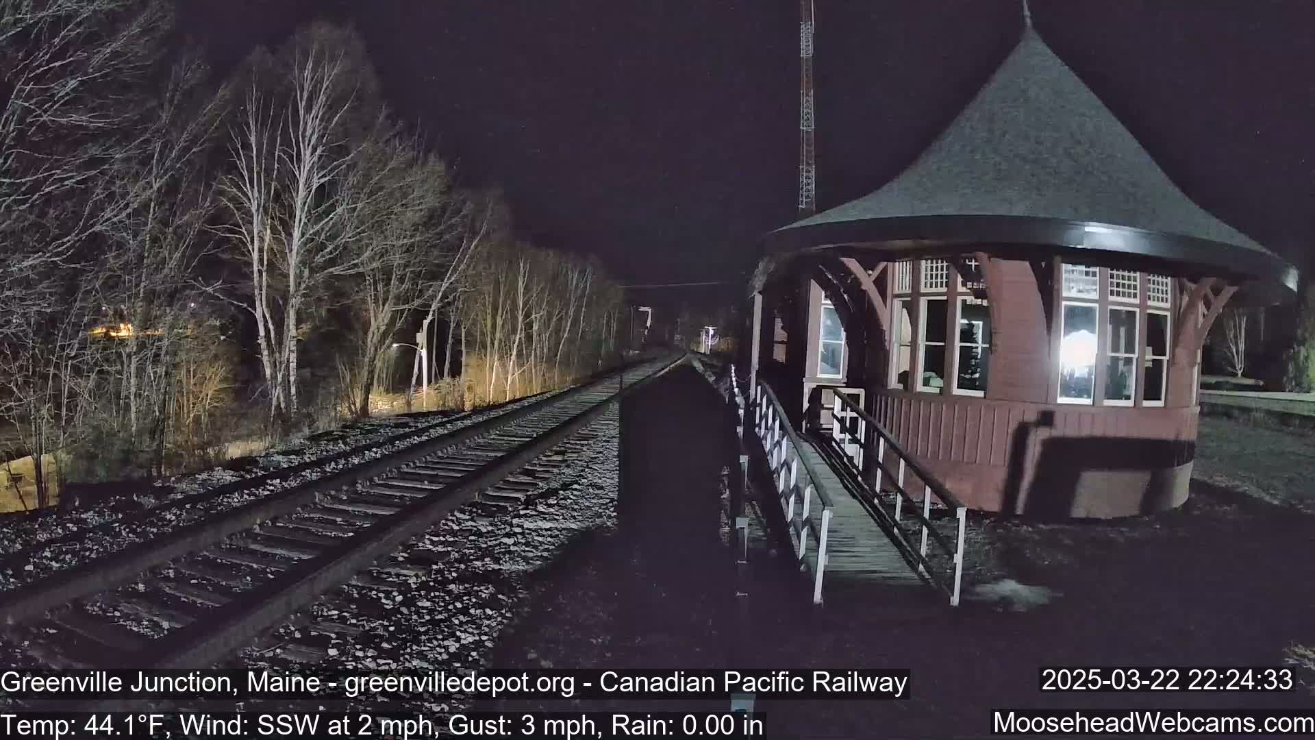 A dark, nighttime scene shows a train station next to railroad tracks, under a lightly raining sky.
