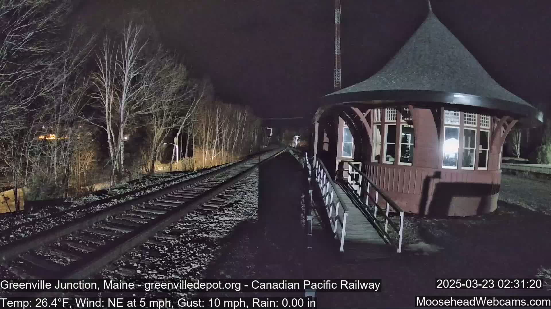 A nighttime view of a train station next to railway tracks under a dark sky with a northeast wind.