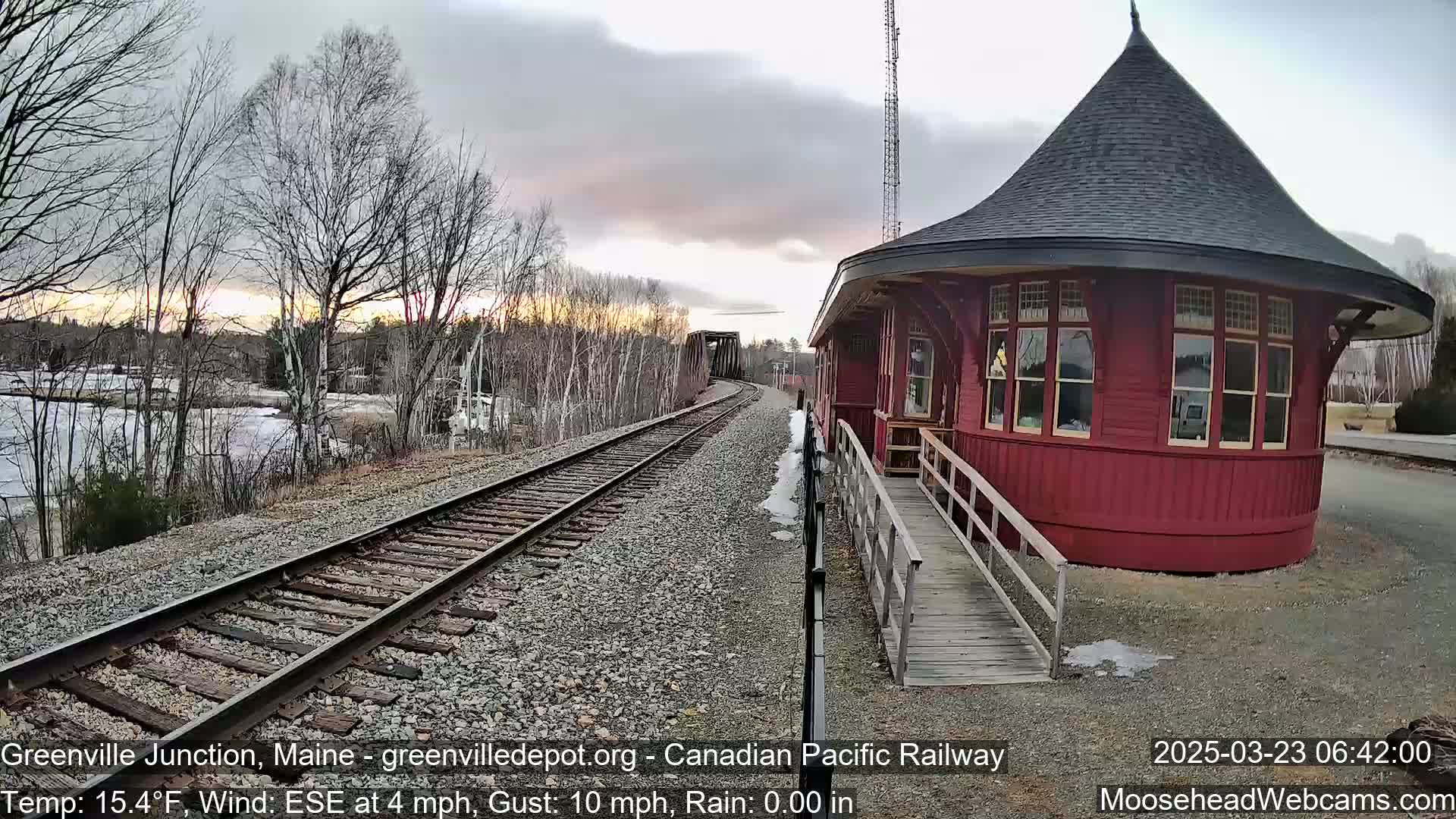A red, round train depot stands beside railroad tracks curving into the distance under a cloudy, sunset sky.