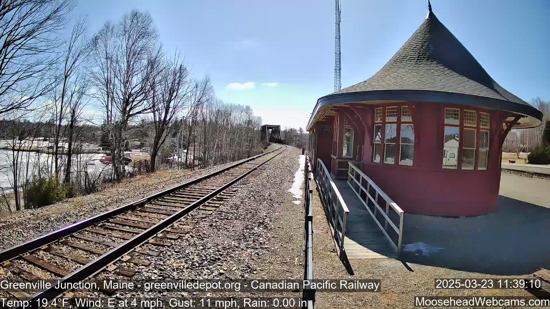 A red, round train station with a conical roof sits beside railroad tracks on a sunny day with a light east wind.
