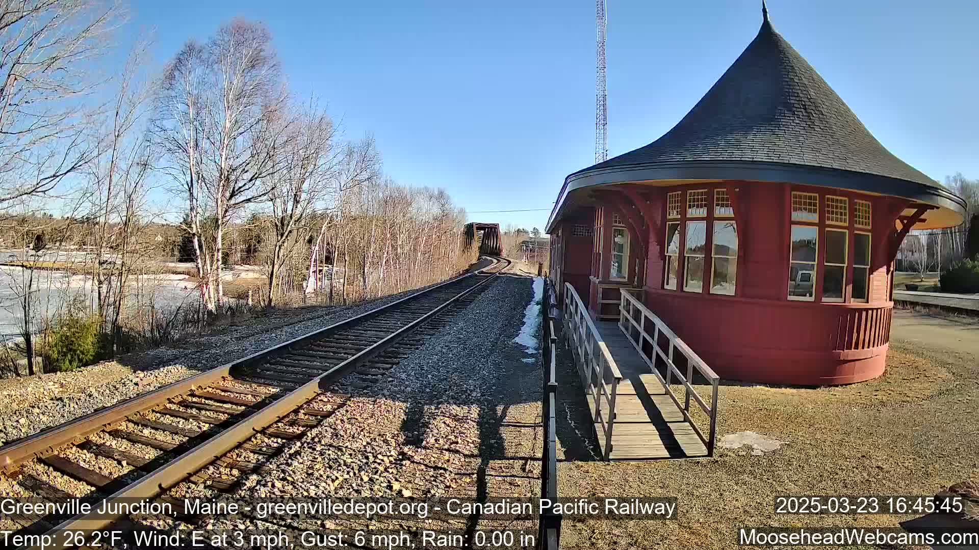 A red, round train depot sits beside railroad tracks leading to a bridge, under a clear blue sky on a sunny, cold day.