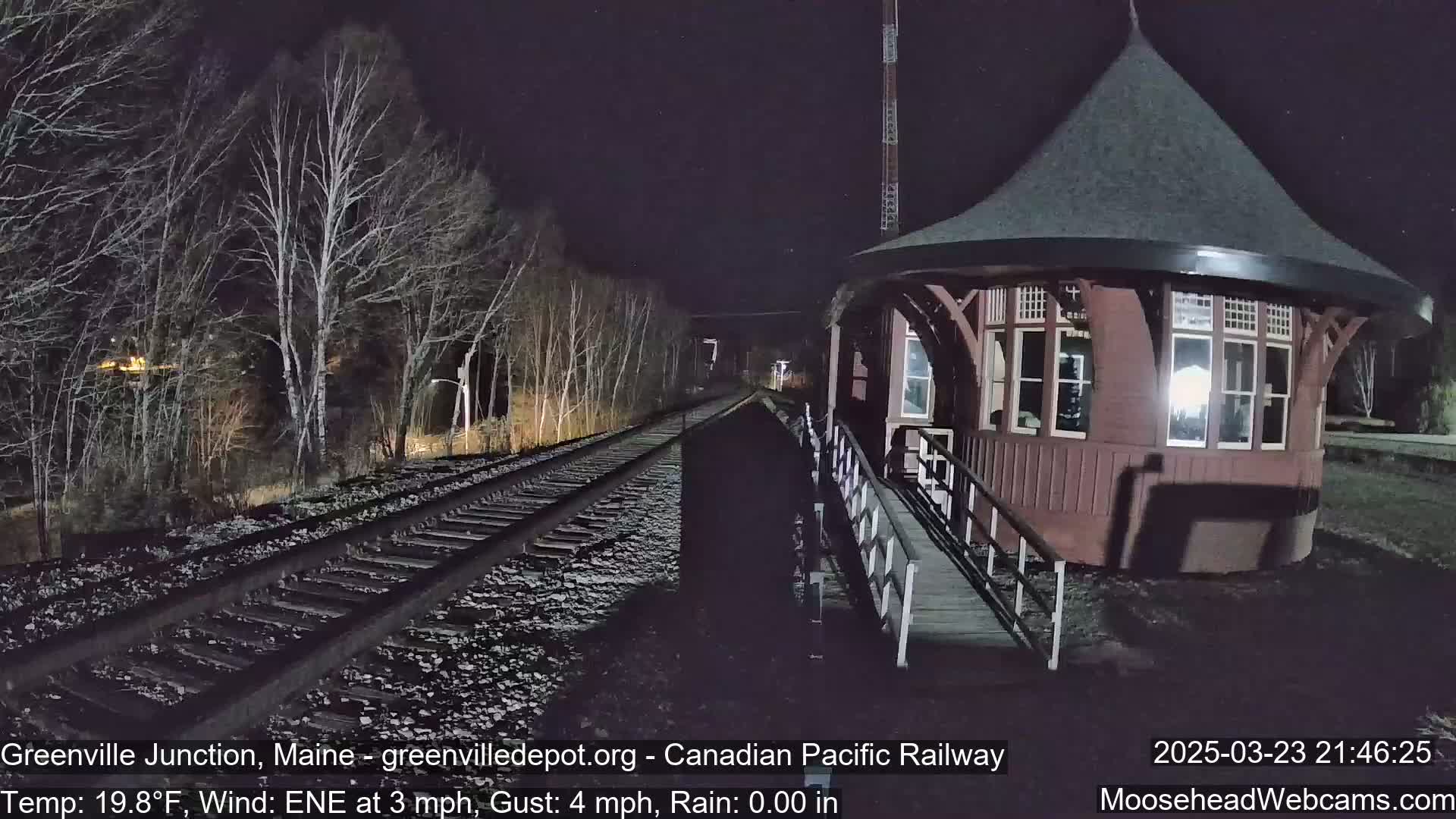 A dark, nighttime scene shows a red octagonal train depot next to railroad tracks lined with bare trees under a clear sky.
