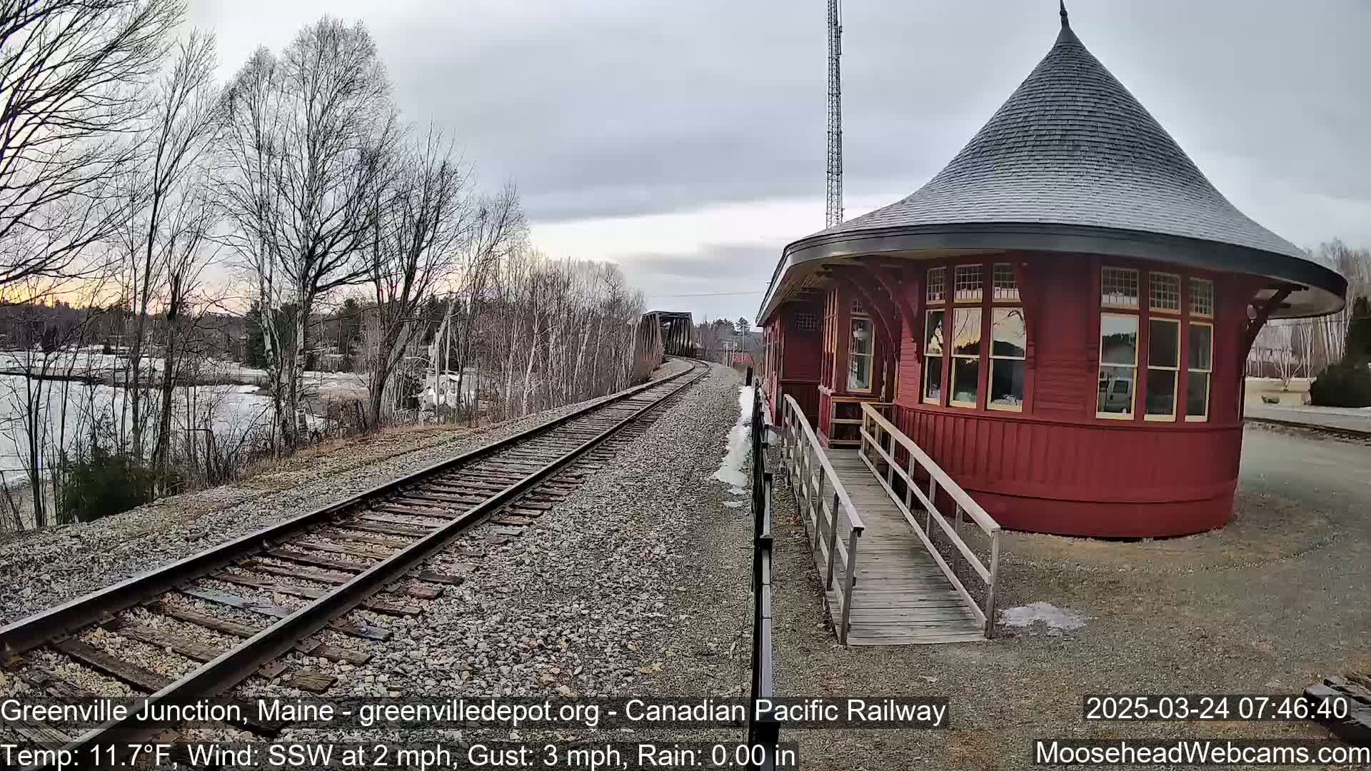 A red, round train depot with a conical roof sits beside train tracks extending to a bridge, under an overcast sky with light rain and bare trees in the background.
