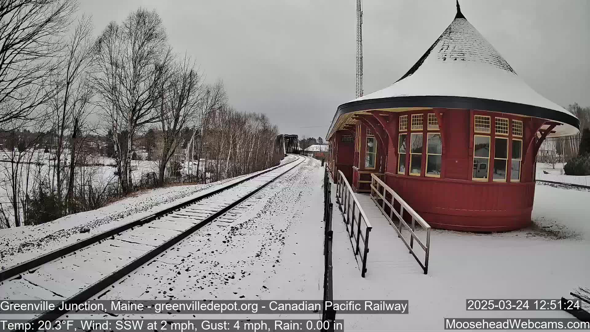 A snow-covered train platform with a red, round train station building is visible next to a railway track on a cold, slightly windy day.