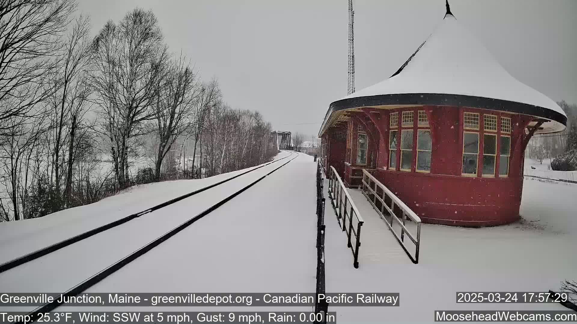Snow falls on a snow-covered train platform next to a red, round train station building.