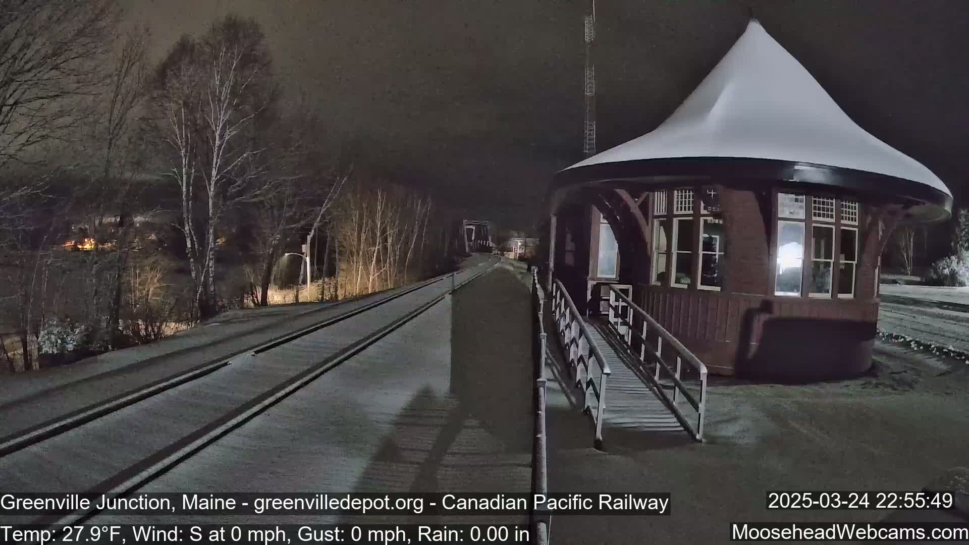 A snow-covered train depot at night, with a gently sloping, snow-covered roof, is situated beside a lightly snow-covered railway track.