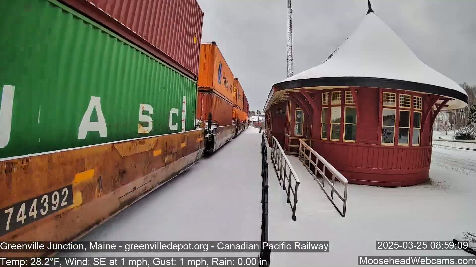 A freight train carrying stacked shipping containers passes a snow-covered, round red train station in snowy weather.