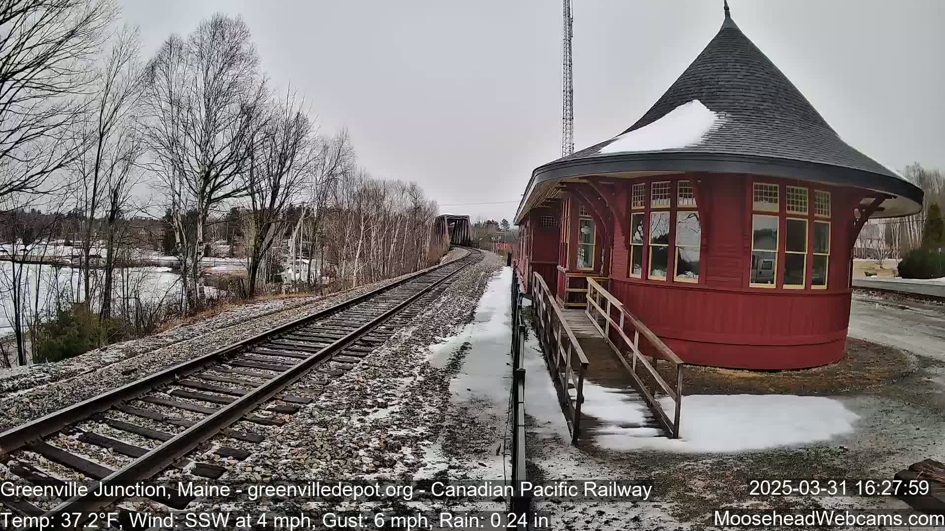 A red, octagonal train depot with a snow-covered roof sits beside railroad tracks, a bridge in the distance, and a partially frozen body of water under a gray, rainy sky.