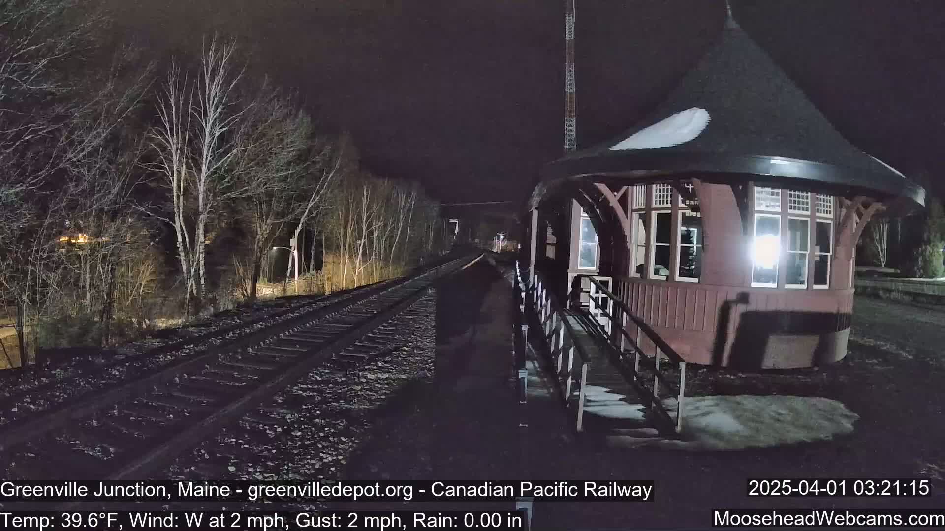 A dark, nighttime scene shows a train station with a snow-dusted roof next to railroad tracks that extend into a wooded area under a clear, dark sky.