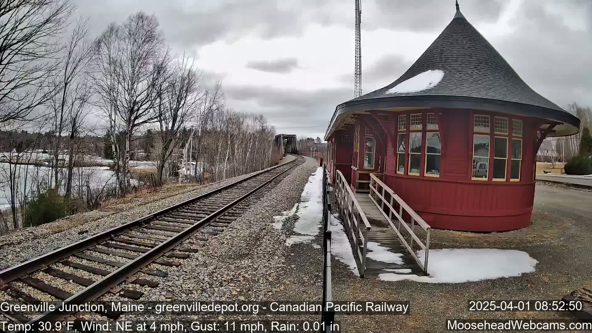 A red, round train station with a conical roof sits beside train tracks on a partly cloudy day with light rain and snow patches on the ground.
