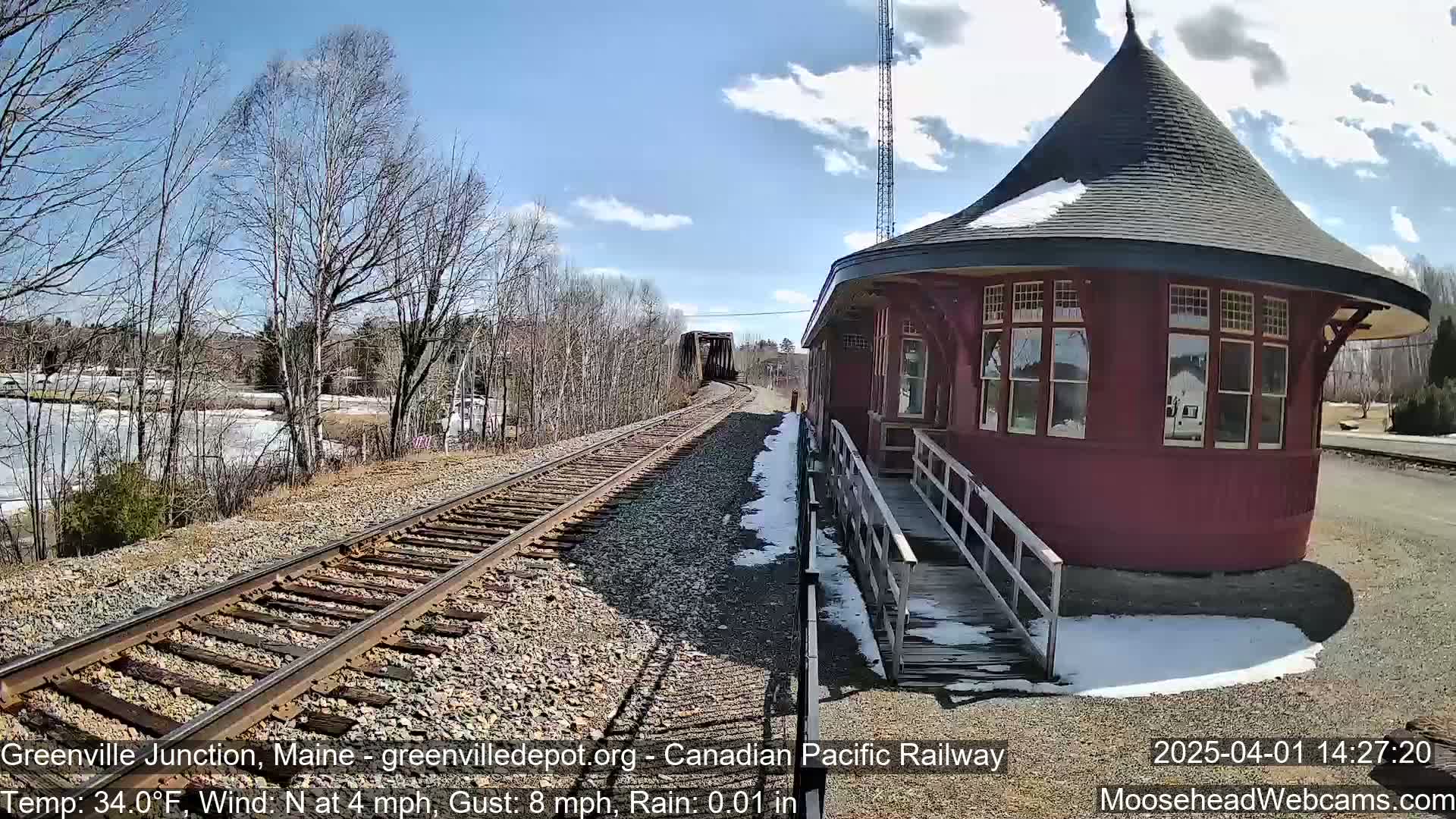 A red octagonal train station with a conical roof stands beside railroad tracks on a partly sunny day with light rain and some remaining snow patches.