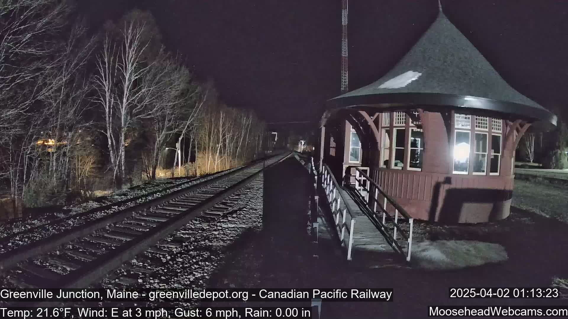 A snow-dusted, octagonal train station stands beside a set of railway tracks at night, under a dark sky with bare trees lining the tracks.