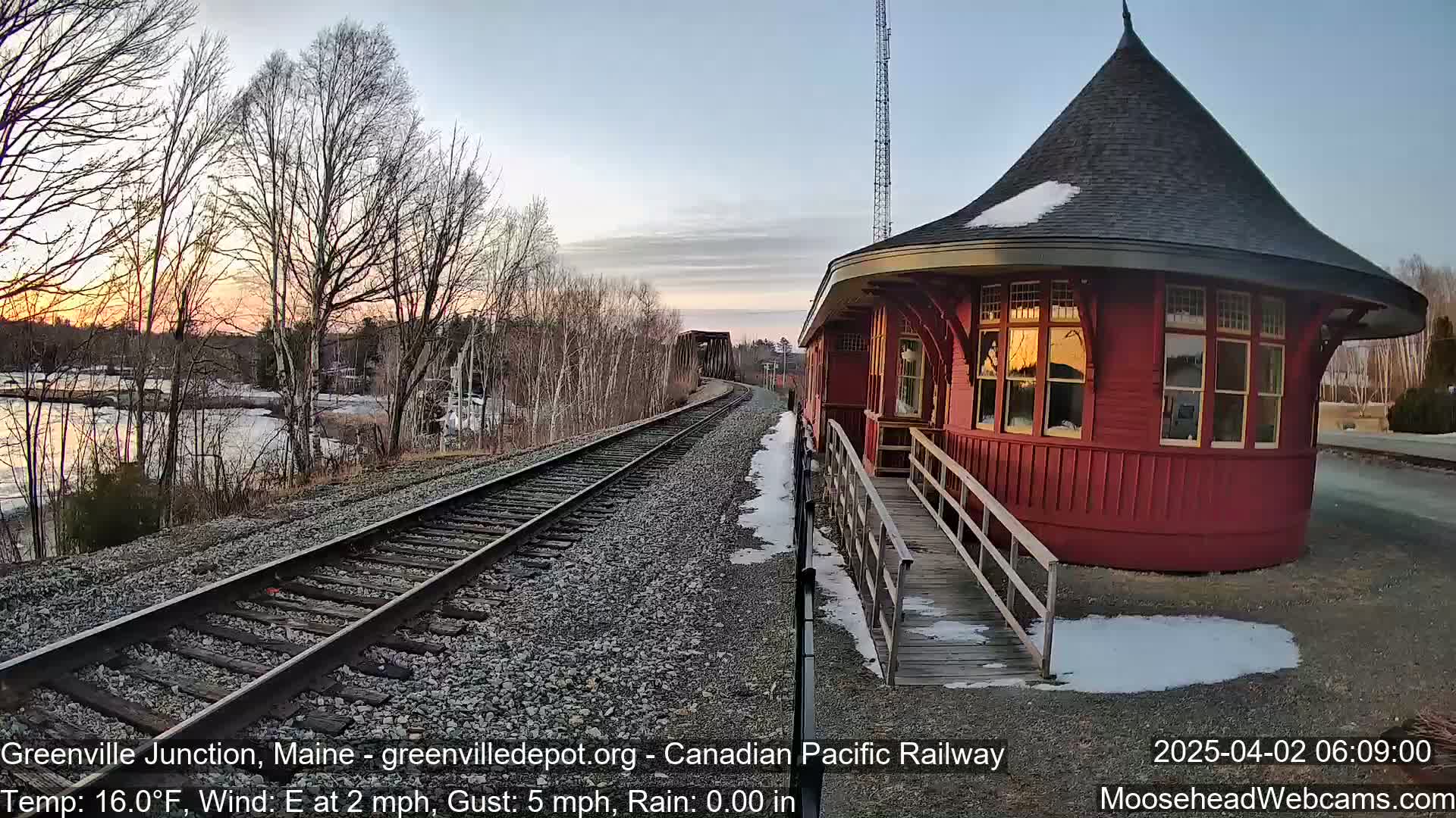 A red, round train station with a ramp sits beside train tracks that lead to a bridge and a calm river under a clear, sunrise sky.