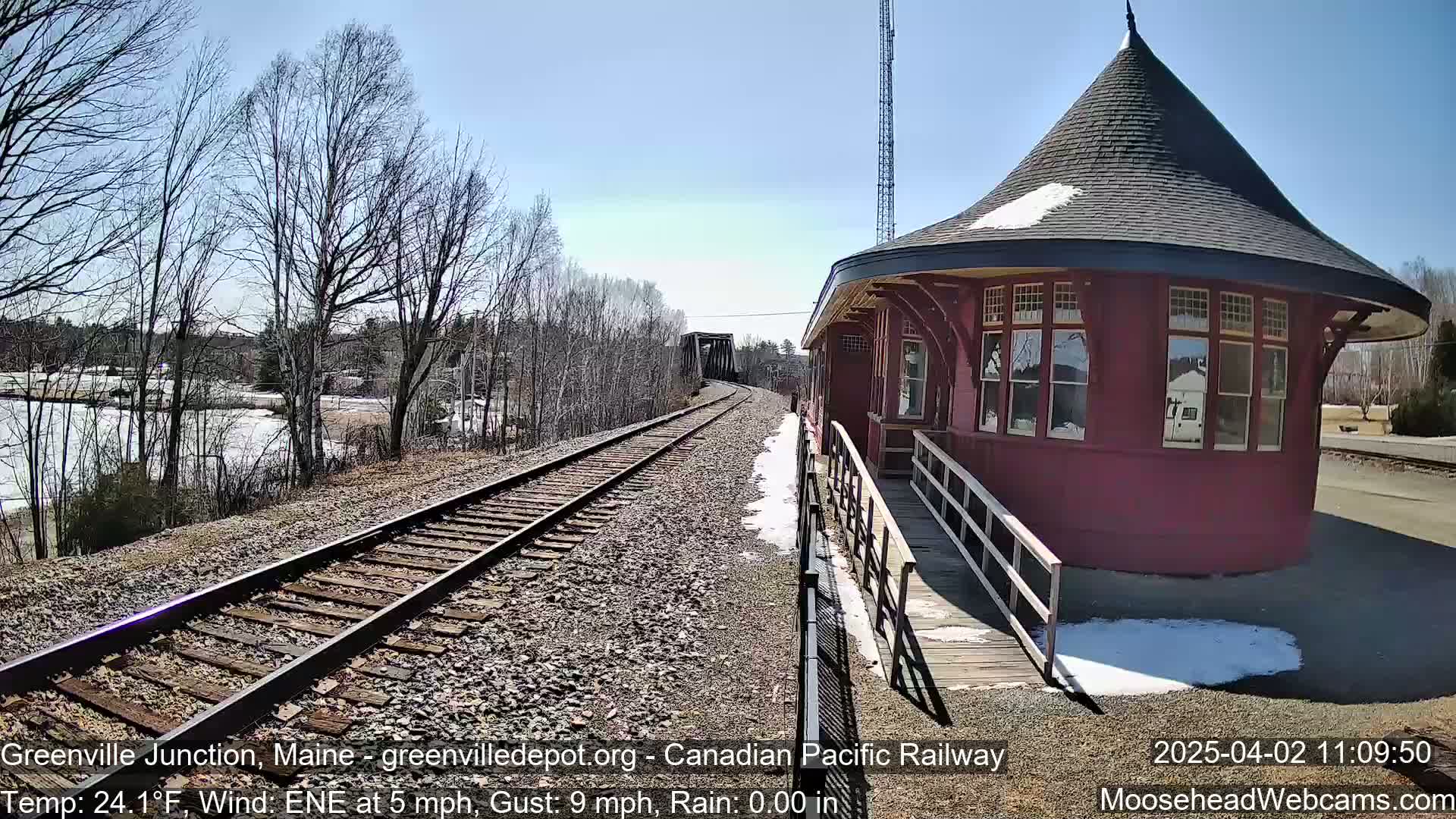 A red, octagonal train station with a conical roof stands next to train tracks on a sunny day with some remaining snow patches and bare trees in the background.