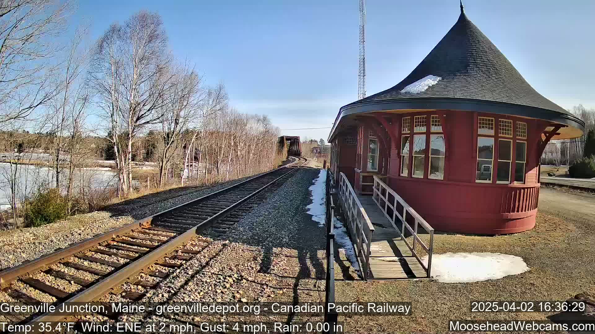A red, round train depot stands beside train tracks on a sunny day with a light wind, a partially snow-covered platform, and a partially frozen body of water in the background.