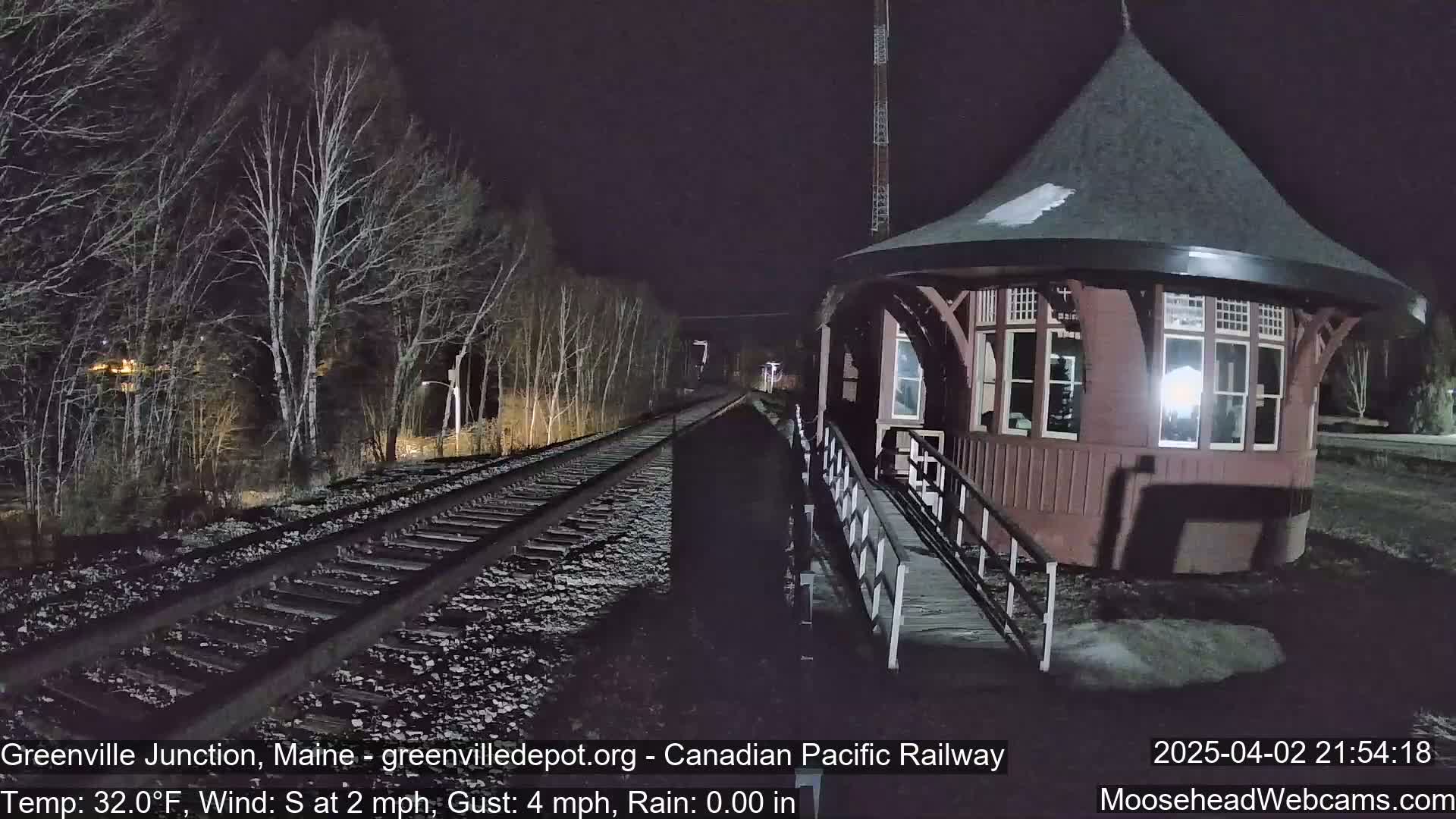 A nighttime view of train tracks leading to a small, red octagonal building beside bare trees under a dark sky.