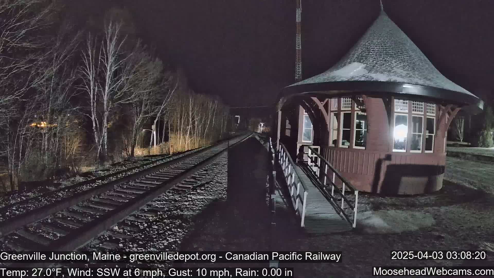 A dark red, octagonal train depot stands next to railroad tracks under a night sky, with bare trees in the background and a light dusting of snow on the ground.