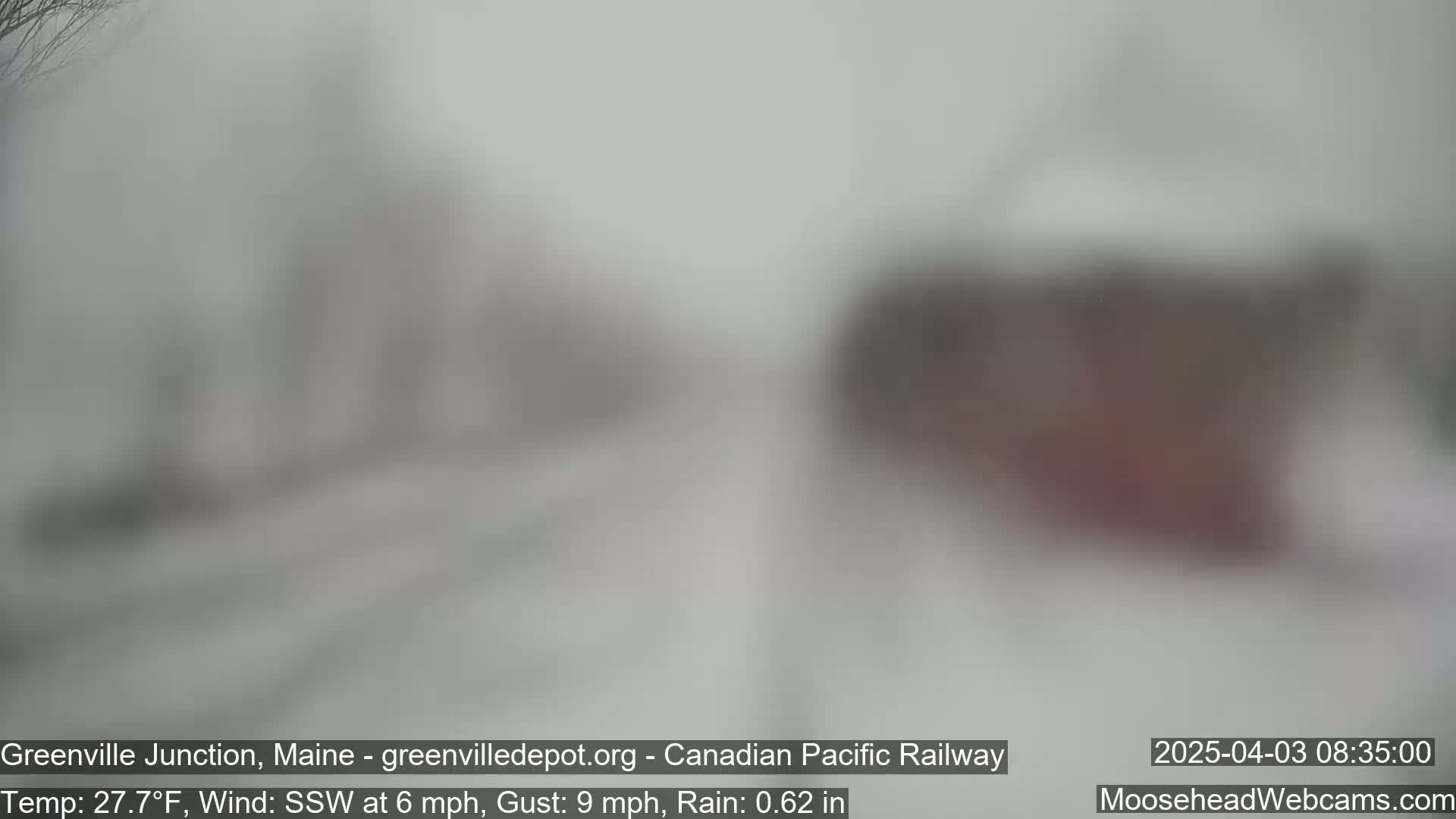 A blurry, snowy outdoor scene shows indistinct buildings and trees under heavy rain and wind.