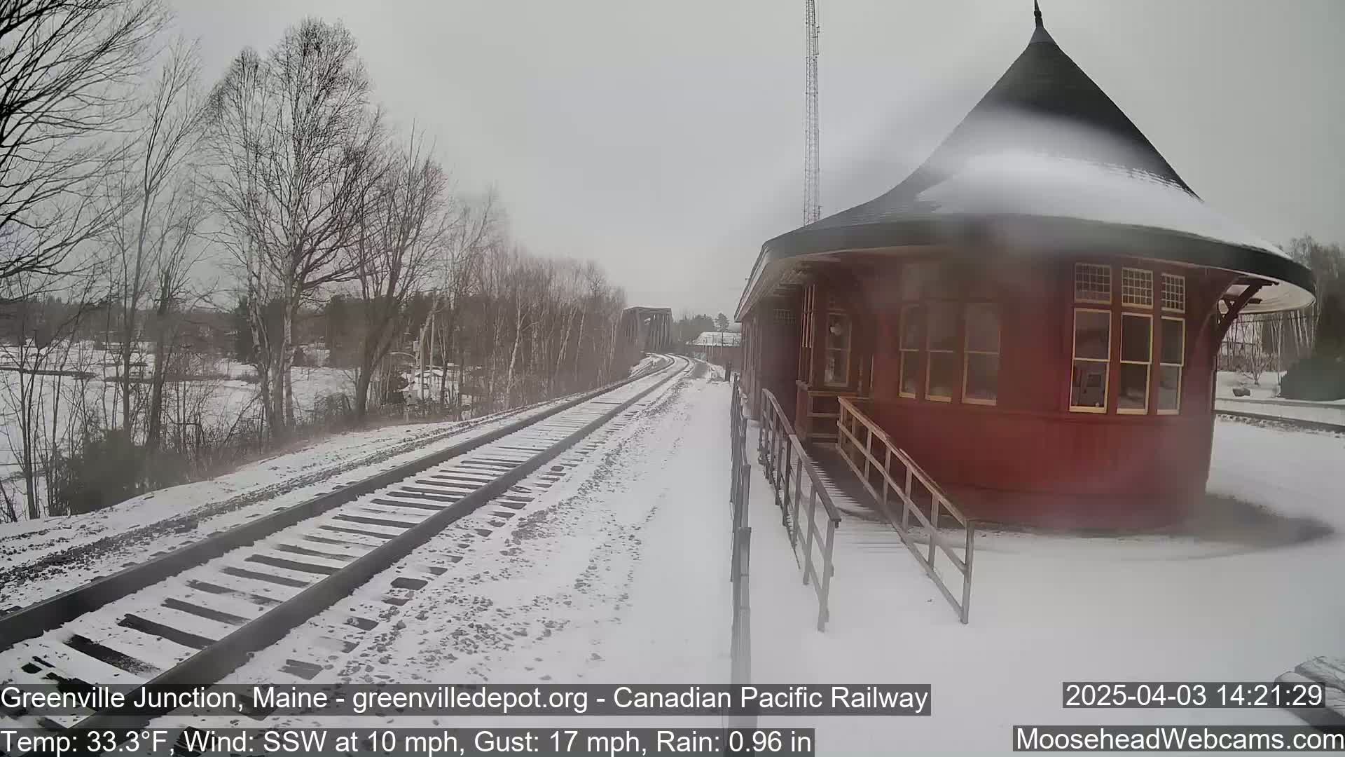 A snow-covered railway track extends toward a red, octagonal train station under a gray sky with light rain and a wind blowing from the south-southwest.