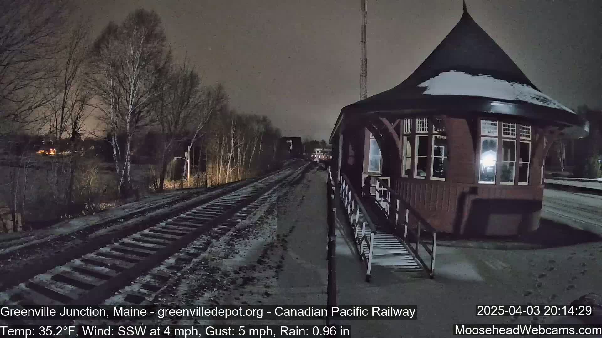 A snow-covered railway track leads to a dark, wooden train station under a nighttime sky with rain.