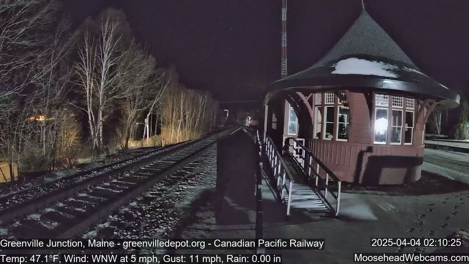 A snow-covered train platform with a red-brown, octagonal building and railroad tracks extends into a dark, sparsely treed landscape at night, under a windy, cool, and rainless sky.