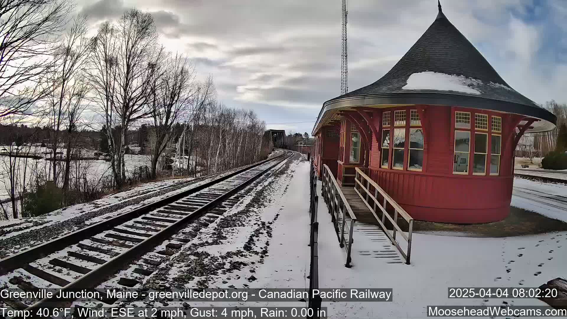 A red, round train station with a conical roof stands beside snow-covered train tracks on a partly cloudy day.