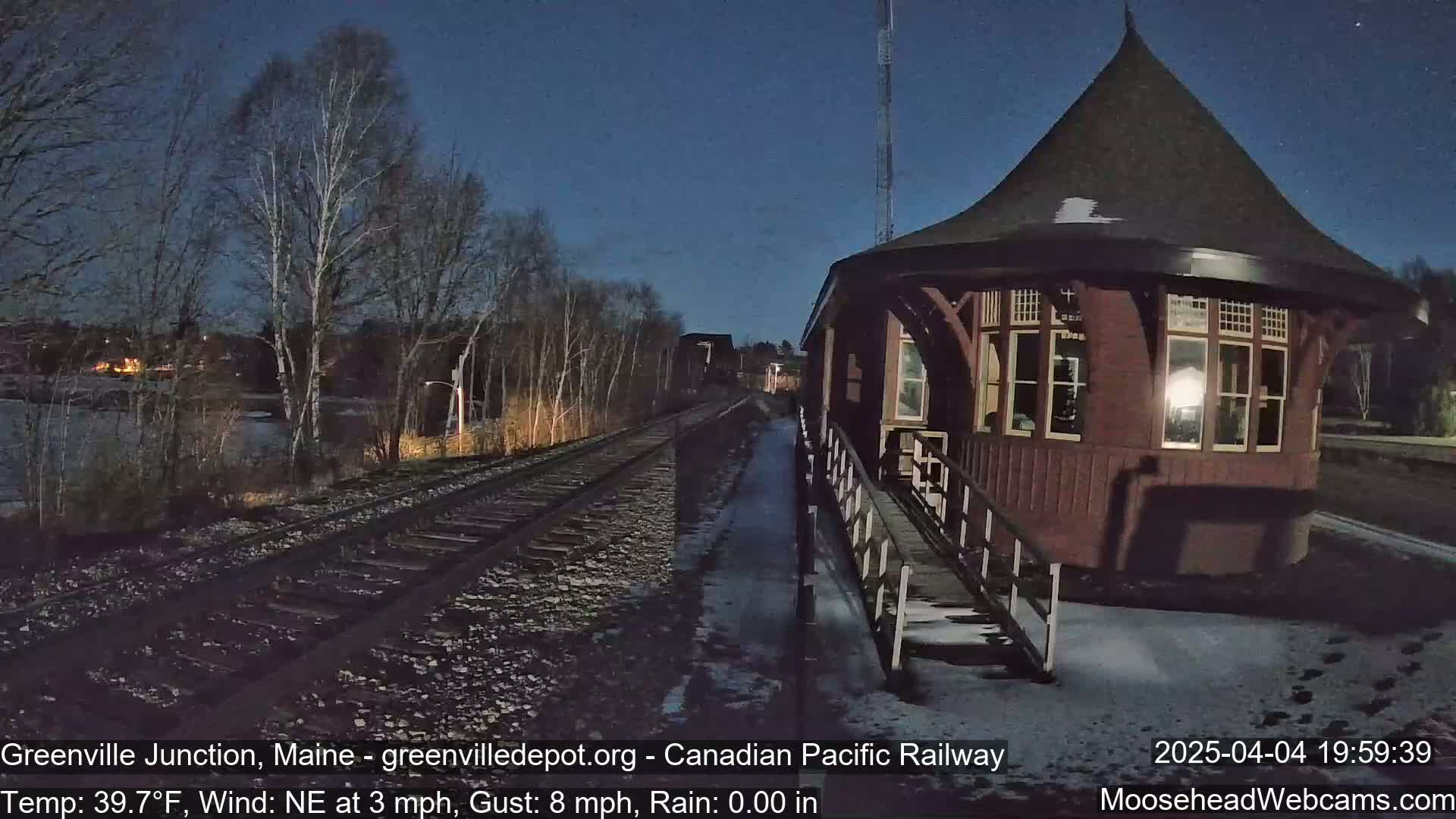 A dark reddish-brown octagonal train depot stands beside railroad tracks under a night sky, with a light dusting of snow on the ground and bare trees in the background.