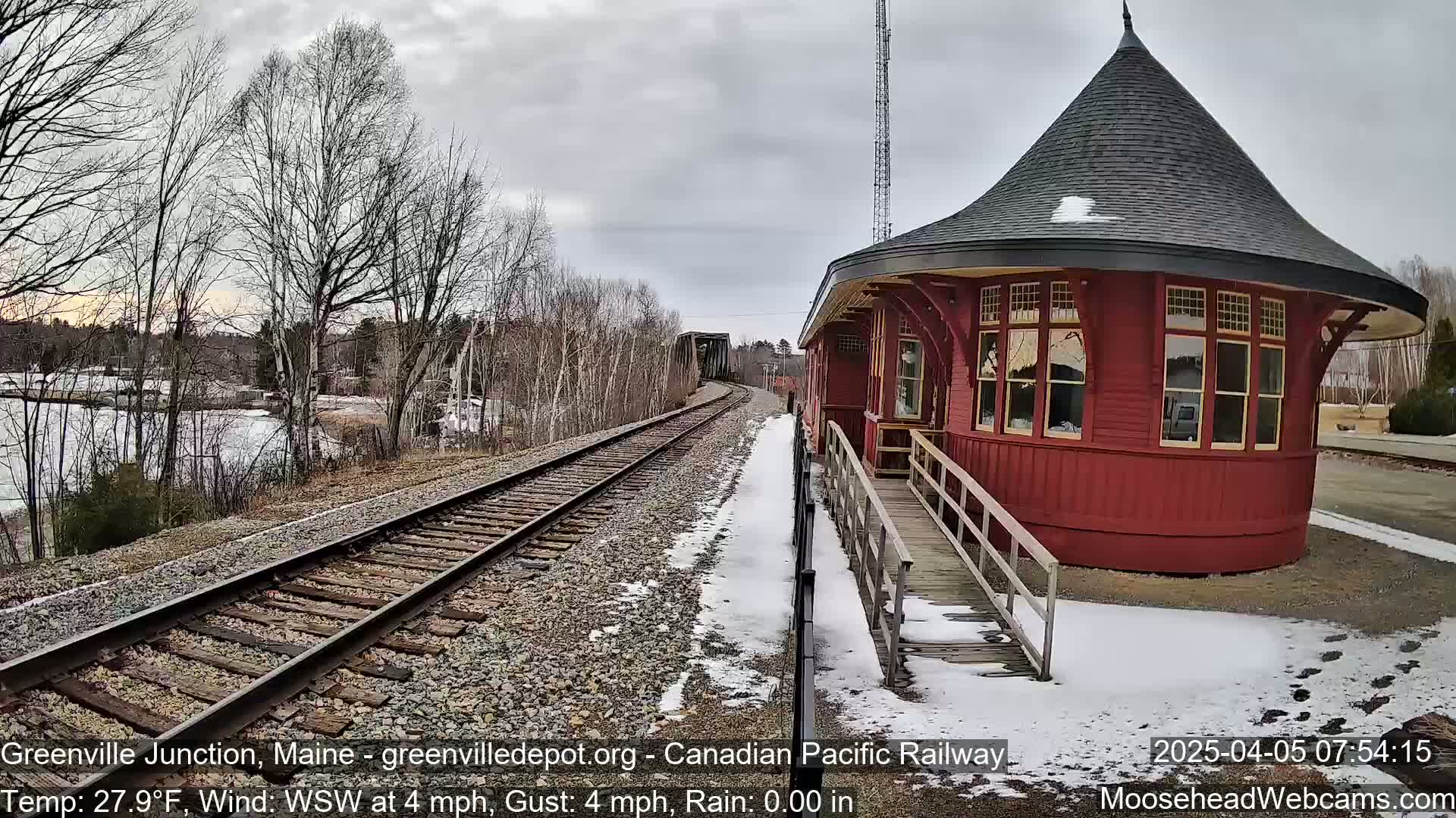 A red, round train station with a conical roof stands beside railroad tracks on a partly snowy, overcast day.