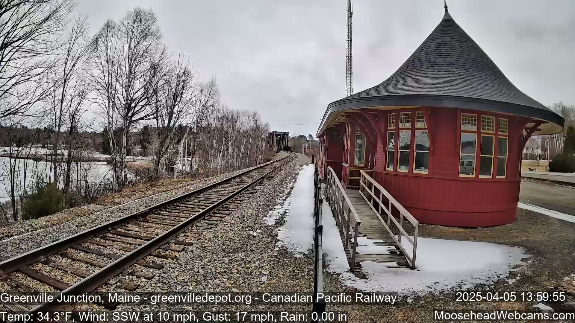 A red, round train depot with a conical roof sits beside railroad tracks that lead to a bridge under a cloudy sky with patchy snow on the ground.