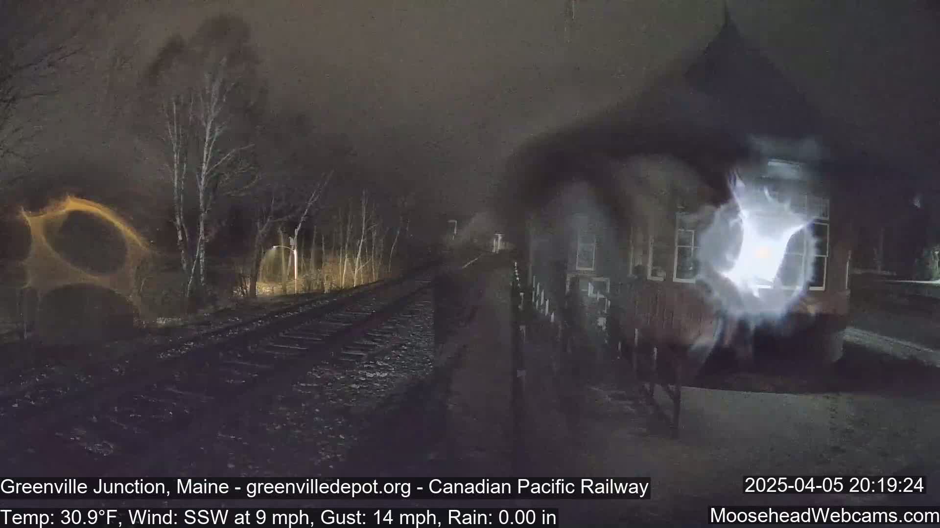 A nighttime view of railroad tracks leading to a building under a dark, rainy sky with a southwest wind.